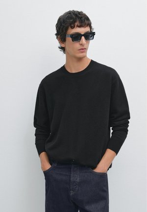 Pull en tricot noir avec un col rond, des manches longues et des poignets côtelés, associé à un jean en denim foncé et des lunettes de soleil noires rectangulaires.