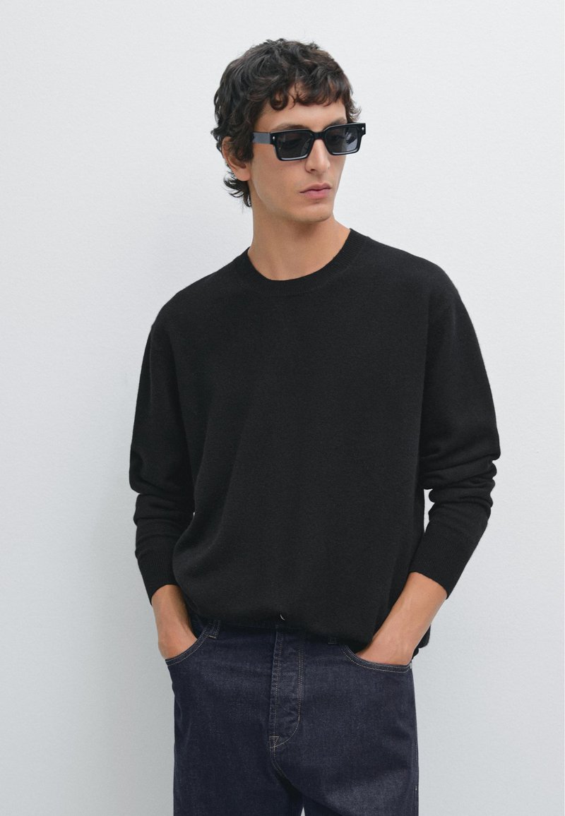 Czarny sweter z dzianiny z okrągłym dekoltem, długimi rękawami i ściągaczami na mankietach, w połączeniu z ciemnoniebieskimi dżinsami i czarnymi prostokątnymi okularami przeciwsłonecznymi.