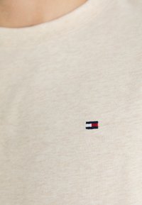 Camiseta de algodón beige con un pequeño logo bordado en rojo, blanco y azul. La tela tiene un aspecto suave y texturizado.
