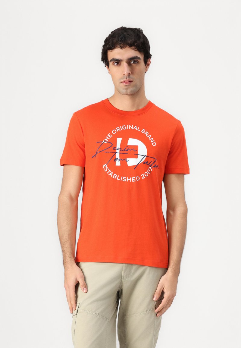 Orange bomulds t-shirt med korte ærmer, rund halsudskæring og grafisk tekst foran i marineblå og hvid. Modell står med hænderne afslappet.