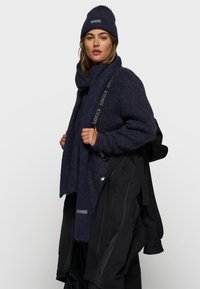 Donkerblauwe gebreide trui en sjaal van fuzzy materiaal, gecombineerd met een bijpassende beanie. Zwarte buitenjas met eenvoudige hardwaredetails.