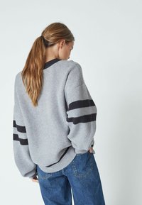 Szürke, oversize pulóver fekete csíkokkal, kötött textúrával, bordás mandzsettákkal és hemvéggel. Magas derékkal ellátott kék farmernadrággal párosítva.