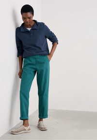 Polo à manches longues bleu marine avec un col, associé à un pantalon droit teal et des sandales tissées beiges.