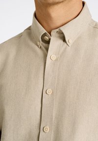 Camisa de botones de manga corta en color beige, confeccionada con una tela texturizada, con un cuello clásico y botones de color claro, que presenta una tapeta delantera limpia.