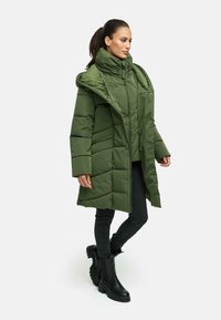 Abrigo puffer verde con cuello alto y cremallera frontal, con paneles acolchados y bolsillos laterales. Combinado con jeans skinny negros y botas de tobillo.