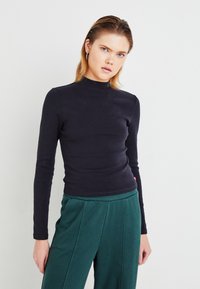 Svart langermet turtleneck-genser i en tettsittende design, laget av elastisk stoff. Båret med grønne vide bukser, som har en myk tekstur.