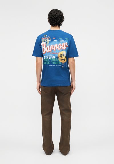 Άνδρας με σγουρά μαλλιά που φοράει μπλε γραφικό μπλουζάκι με πολύχρωμο κείμενο και emoji και καφέ παντελόνι, στέκεται γυρισμένος πλάτη σε λευκό φόντο.