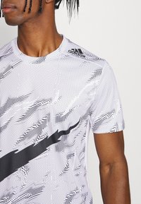 Camiseta atlética blanca con un patrón abstracto negro y un logo. Presenta un cuello redondo y mangas cortas, confeccionada en una tela suave y ajustada.