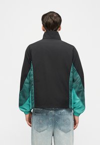 Un homme porte une veste noire et bleu sarcelle avec des manches à motifs et un jean bleu clair présentant des broderies sur les poches arrière.