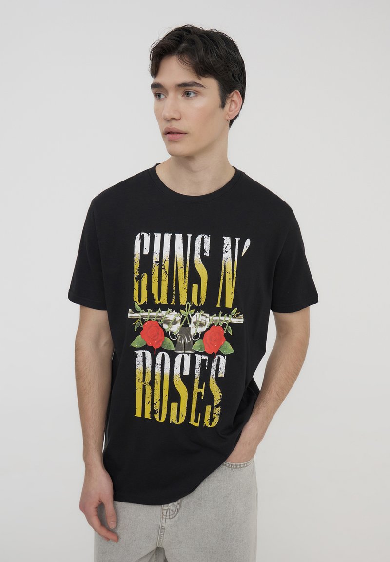 T-shirt in cotone nero con la scritta "GUNS N' ROSES" in giallo invecchiato, accompagnata da un grafico di revolver e rose rosse sul davanti.
