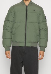 Groene gewatteerde jas met een ritssluiting aan de voorkant, geribde manchetten en zoom. Heeft een logo op de borst en een zijzak aan de linkerkant.