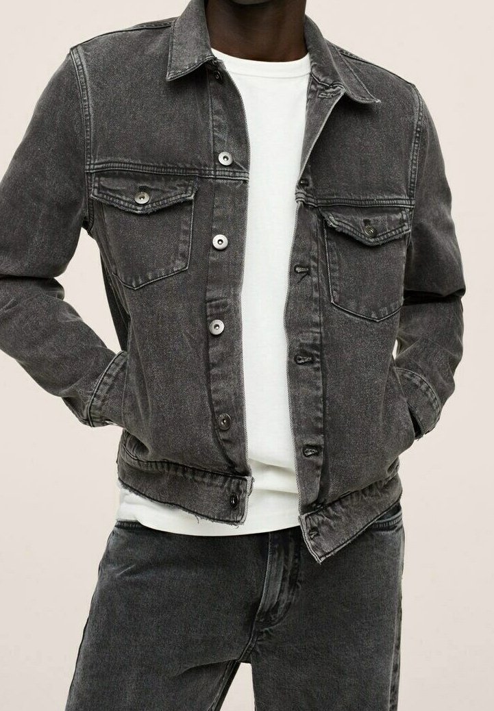 Uomo che indossa una giacca di jeans nera sbiadita sopra una camicia bianca, con le mani nelle tasche della giacca, abbinata a jeans di jeans neri.