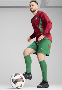 Rote und grüne Fußballausrüstung mit langen Ärmeln und Shorts, die Muster und das Puma-Logo zeigt. Schwarze Fußballschuhe und ein mehrfarbiger Ball.