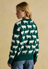 Grüner Strickpullover mit einem sich wiederholenden weißen Schafmuster, langen Ärmeln und einem runden Halsausschnitt. Weiche Textur und lockere Passform.