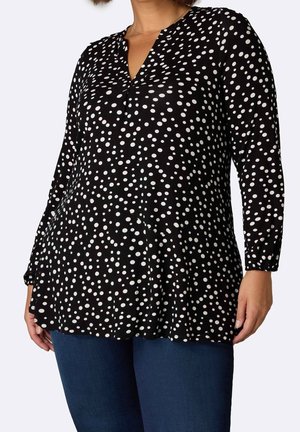 Frau trägt eine schwarze Langarm-V-Ausschnitt-Bluse mit weißen, unregelmäßigen Punkten und blaue Jeans, steht vor einem schlichten Hintergrund.