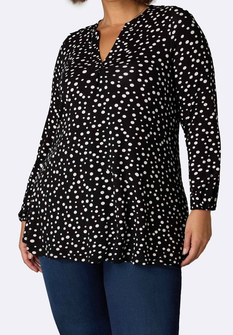 Frau trägt eine schwarze Langarm-V-Ausschnitt-Bluse mit weißen, unregelmäßigen Punkten und blaue Jeans, steht vor einem schlichten Hintergrund.