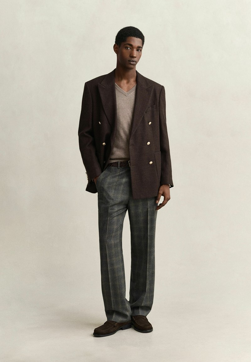 Joven de pie que lleva un blazer cruzado marrón oscuro, un suéter beige con cuello en V, pantalones grises de cuadros y zapatos marrones.