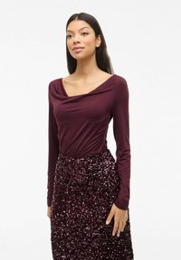 Bordeaux lang mouwtop met een gedrapeerde halslijn, gecombineerd met een sequined bordeaux rok. Gladde textuur op de top; glanzende, gestructureerde sequins op de rok.