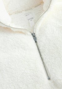 Hvid fleece-pullover med kvart lynlås, der har blød tekstur og sølv hardware. Etiketten indeni læser "downtime NEXT."