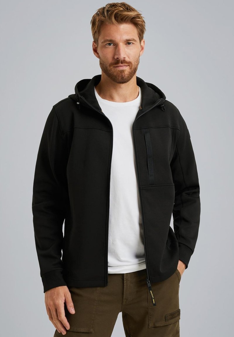 Sudadera negra con cremallera en el frente, capucha ajustable, bolsillo en el pecho con cremallera y puños de canalé, usada sobre una camiseta blanca. Textura suave, ajuste casual.