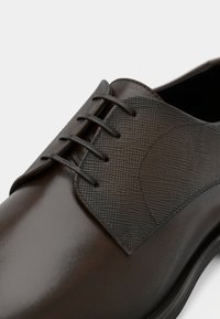 Brauner Lederschuh mit texturierter dunkelbrauner Fläche, fünf Ösen und glatter Oberfläche. Runde Spitze und dezente Nähte.