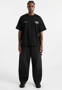 Czarna bawełniana koszulka z krótkim rękawem i luźnym krojem. Na przodzie znajduje się żółty nadruk graficzny i tekst. Noszona z luźnymi czarnymi dżinsami.