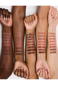 Quatre bras présentent des échantillons de rouge à lèvres dans différentes nuances de nude et marron. Des étiquettes indiquent le nom de chaque teinte sur le deuxième bras en partant de la gauche.