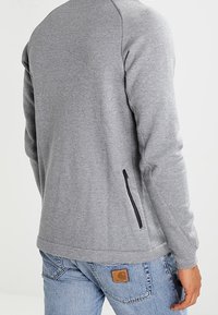 Harmaa huppari, jossa on silkkinen pinta, mustalla vetoketjullisella taskulla sivussa ja ribbireunailla, käytetty sinisten farkkujen kanssa.