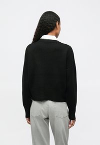 Γυναίκα με μακριά κυματιστά μαλλιά, ντυμένη με μαύρο πουλόβερ πάνω από λευκό πουκάμισο και ανοιχτό γκρι τζιν, στέκεται με την πλάτη στραμμένη προς την κάμερα σε λευκό φόντο.