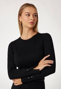 Haut noir en tricot à manches longues avec un col rond et des détails de boutons sur les poignets. Texture lisse, design ajusté.