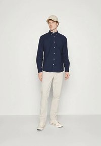 Marineblå skjorte med hvite knapper, kombinert med lysebeige bukser og beige caps. Modellen står mot en ensfarget hvit bakgrunn.