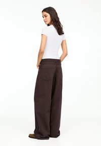 Tricou alb ajustat, asociat cu pantaloni largi, de culoare maro închis, cu talie înaltă și buzunare pe spate vizibile prin cusături.