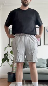 Pantaloni shorts grigi chiari con cuciture laterali e tasche, dotati di una fascia elastica in vita. Abbinati a una t-shirt nera e calze sportive bianche.
