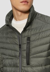 Leichte olivgrüne Pufferjacke mit horizontaler Steppstruktur, Reißverschluss und einem schwarzen Markenband auf der Brust.