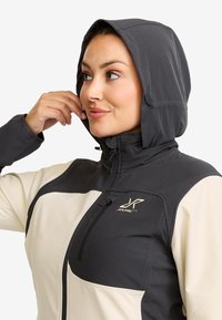 Woman adjusting the hood of a two-tone black and beige zip-up jacket with a chest pocket and RVRC logo.