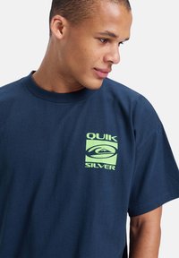 Mørkeblå bomull t-skjorte med en limes grønn logo med "QUIK SILVER" tekst og fjelldesign på venstre brystområde. Korte ermer.