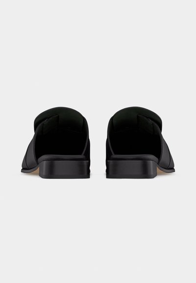 Ζευγάρι μαύρα δερμάτινα μοκασίνια χωρίς κορδόνια με χαμηλό τακούνι, εμφανιζόμενα από πίσω σε λευκό φόντο.