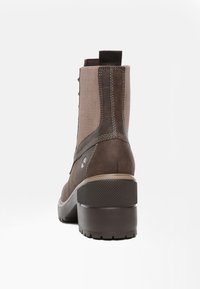 Botín de tobillo de cuero marrón y tela con tirador, tacón grueso texturizado y detalles cosidos, mostrado desde atrás sobre fondo blanco.