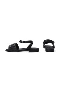 Schwarze Sandalen mit einer strukturierten Oberseite, verstellbarem Knöchelriemen und flacher Sohle. Auffällige Hardware-Akzente am vorderen Riemen.