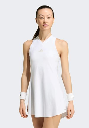 Mujer vistiendo un vestido de tenis blanco sin mangas con el logo de Adidas, acompañado de muñequeras blancas, de pie frente a un fondo claro y liso.