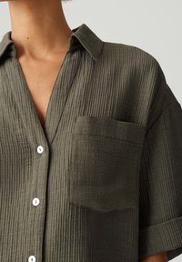Chemise à manches courtes couleur vert olive en tissu texturé à rayures verticales, avec une poche avant et des boutons blancs.