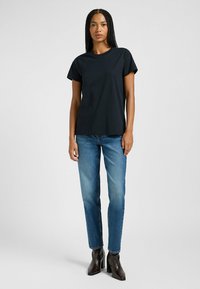 T-shirt nera in cotone con maniche corte, colletto a giro, abbinata a jeans blu e stivaletti neri con tacco, in piedi contro uno sfondo chiaro.