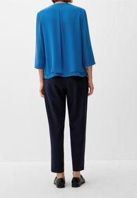 Personne debout, vue de dos, portant un chemisier bleu à manches trois-quarts, un pantalon noir sur mesure et des chaussures noires sur un fond blanc.