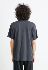 Person med krøllet hår iført en løs sort t-shirt og sorte bukser, stående med ryggen til mod en ensfarvet hvid baggrund.