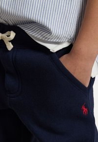 Barn som bär marinblå svettbyxor med röd logotyp och handen i fickan, matchat med en vit och blårandig tröja.