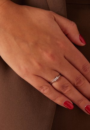 Silver ring with three diamond-like accents, set in a delicate band. Hand placed on a brown textured surface, highlighting the jewelry.