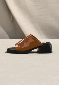 Καφέ λουστρίνι mule με ανοιχτά δάχτυλα, μαύρο χοντρό μεσαίο τακούνι και επίπεδη σόλα σε ουδέτερο υφασμάτινο φόντο.