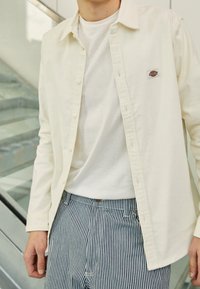 Camisa de botones color beige claro sobre una camiseta blanca, combinada con pantalones de rayas azules y blancas. La tela parece suave y el ajuste es relajado.
