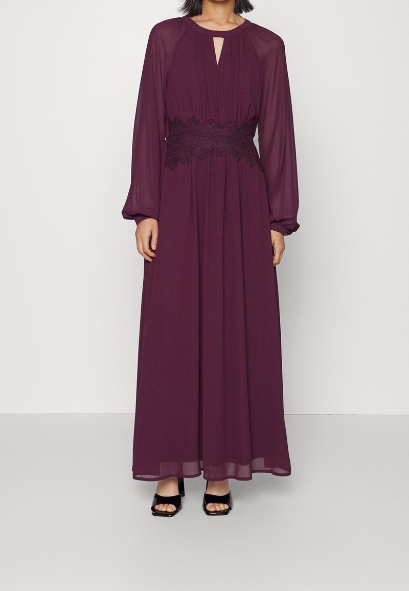 Femme portant une robe longue à manches longues, de couleur violet foncé, avec un détail en dentelle à la taille et des chaussures noires à talons blocs ouvertes à l'avant.