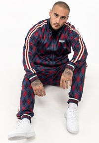 Marineblauwe en rode argyle trainingsuit met witte accenten, ritsjack, elastische manchetten en bijpassende broek, gecombineerd met witte sneakers.